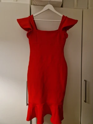 Röd volangklänning med statementärmar - Snygg röd klänning med figurnära passform, fyrkantig urringning och stora statementärmar. Klänningen har volangdetalj nertill och dragkedja i ryggen. Perfekt för dig som vill sticka ut med en färgstark look.