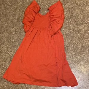 Orange klänning med volang från H&M - Säljer en orange klänning från H&M i storlek 134/140. Klänningen har breda volangärmar och en mjuk, luftig passform. Har inga synliga defekter. Kom gärna med prisförslag!