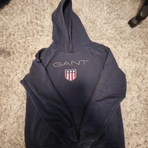 Marinblå hoodie från GANT - Säljer en marinblå hoodie från GANT med broderad logga och sköld på bröstet. Tröjan har huva och en klassisk känguruficka framtill. Perfekt för dig som gillar sportig och avslappnad stil.