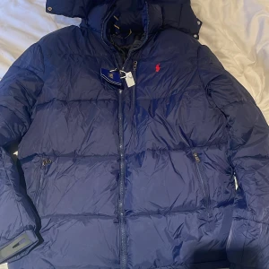 Marinblå pufferjacka från Polo Ralph Lauren - Snygg marinblå pufferjacka från Polo Ralph Lauren med röd logga på bröstet. Jackan är i storlek L och är perfekt för vintern. Den är helt oanvänd endast testad en gång. 