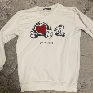 Vit Palm Angels tröja med nalletryck - Säljer en vit långärmad tröja från Palm Angels med ikoniskt nalletryck på bröstet och ett rött hjärta i tyg. Tröjan har rund halsringning och svart logga framtill. Perfekt för dig som gillar streetwear och vill sticka ut med en unik design.