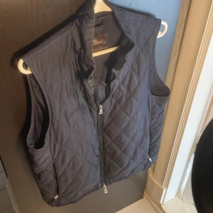 Mörkblå väst Oscar Jacobson  - Quiltad mörkblå väst från Oscar Jacobson med hög krage och dragkedja framtill. Västen har diskret rutmönster, två fickor med dragkedja och en stilren look. Perfekt för lager-på-lager under kyliga dagar. Västen är i toppen skick. Nypris 2600. Kom med bud!