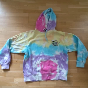 Vans tie-dye hoodie med huva - Färgglad hoodie från Vans i tie-dye med nyanser av rosa, blått, gult och lila. Stor huva med snörning, magficka och tryckt Vans-logga på bröstet. Perfekt för dig som gillar streetstyle och vill sticka ut med en unik look.