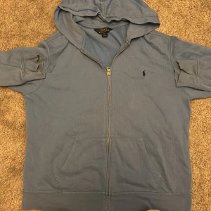 Blå hoodie från Polo Ralph Lauren - Säljer en blå hoodie från Polo Ralph Lauren i storlek XL. Tröjan har dragkedja, huva och känguruficka framtill. Klassisk broderad logga på bröstet och mjukt bomullsmaterial. Perfekt för chill dagar eller när du vill ha en enkel och snygg look.