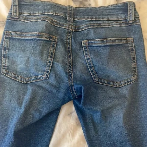 Blå jeans från Gina Tricot, storlek 170 - Snygga blå jeans från Gina Tricot med klassisk femficksdesign och dubbla silverknappar i midjan. Jeansen har en rak passform och är tillverkade i slitstarkt denimtyg med tydliga sömmar. Perfekta för dig som gillar en enkel och tidlös look.