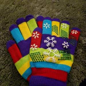 Färgglada randiga stickade vantar - Säljer ett par stickade vantar i flera färger med randigt mönster. Vantarna har snöflingor i vitt på ovansidan och halkskydd i grönt på handflatan. Färgerna är blå, röd, gul, grön, lila och turkos. Perfekta för vintern och riktigt lekfulla i stilen.