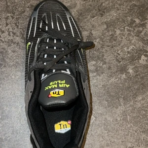 Nike Air Max Plus svart/grå sneakers - Nike Air Max Plus sneakers i svart och grått med limegröna detaljer och TN Air-logga. Ovandel i mesh och syntet med snörning, samt synlig Air Max-dämpning i sulan. Snyggt randigt mönster och klassisk rund tå. Perfekt för dig som gillar streetwear och sportig stil.