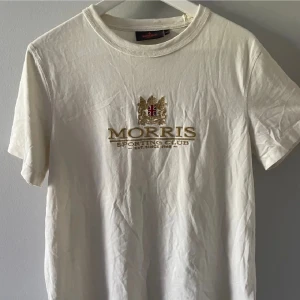 Vit t-shirt från Morris Sporting Club - Vit t-shirt från Morris Sporting Club med broderad logga och gulddetaljer framtill. Klassisk rund halsringning och korta ärmar. Skön och stilren modell i bomull, perfekt för en clean och sportig look.