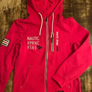 Cericeröd hoodie från Nautic, stl 36 - Riktigt snygg röd hoodie från Nautic XPRNC RS65 med broderad logga och text framtill. Tröjan har huva med snörning, dragkedja och två fickor. Detaljerad patch på ärmen och tillverkad i ekologisk bomull. Perfekt för dig som gillar sportig och avslappnad stil.