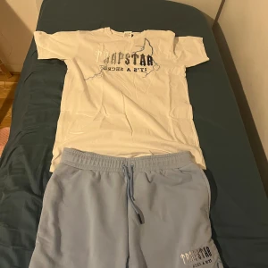 Vit Trapstar t-shirt med tryck - Vit t-shirt, Trapstar med stort tryck och broderade blixtdetaljer på bröstet. Snyggt tryck i silver och svart med texten 'Trapstar It's a Secret'. Klassisk passform och rund halsringning. Perfekt för dig som vill sticka ut med streetwear-vibe.