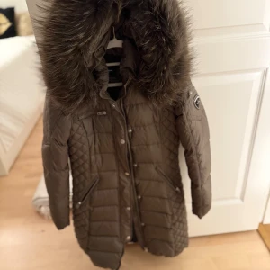 Brun lång pufferjacka med fuskpäls - Snygg brun pufferjacka från Rockandblue med lång passform, quiltade detaljer och stor huva med fuskpäls. Jackan har dragkedjor på fickorna, tryckknappar framtill och en patch på ärmen. Perfekt för kalla dagar och riktigt mysig att bära.