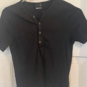 Svart ribbad topp från Gina Tricot - Svart ribbad topp från Gina Tricot med korta ärmar och knappdetaljer framtill. Snygg och enkel design som passar till många olika stilar. Mjuk bomullskvalitet och slim passform för en skön känsla.