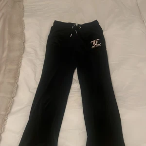 Svarta mjukisbyxor Juicy Couture - Svarta mjukisbyxor från Juicy Couture med broderad logga framtill. Byxorna har snörning i midjan, raka ben och bakfickor med silverfärgade knappar. Mjukt material som känns lyxigt och skönt mot huden. Inte alls mycket andvända