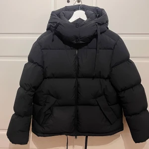 Svart pufferjacka från GANT - Svart pufferjacka från GANT med huva. Perfekt för kalla dagar. Köpt på NK 2024 och jackan är i nytt skicka med inga skador! Kan mötas i Stockholm 