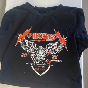 Svart croppad t-shirt Pursuers - Svart croppad t-shirt från Pursuers med tryck av örn, blixtar och text i rött och vitt. T-shirten har korta ärmar och rund halsringning. Perfekt för dig som gillar en edgy och sportig stil.