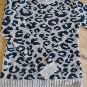 Beige leopardmönstrad stickad tröja - Säljer en beige stickad tröja från NA-KD med leopardmönster i svart och brunt. Tröjan har rund halsringning, ribbade muddar och en loose passform. Perfekt för dig som vill sticka ut med en trendig look.