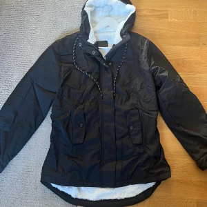 Svart parkajacka med fuskpälsfoder - Svart parkajacka från H&M med vit fuskpälsfoder i huvan och insidan. Jackan har dragsko i midjan, två stora fickor med knappar och dragkedja framtill. Perfekt för kyliga dagar med sin långa passform och mysiga material.