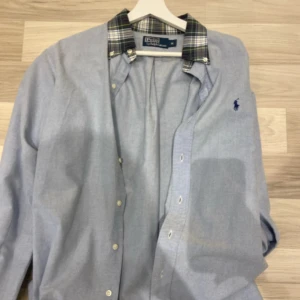 Blå skjorta Polo Ralph Lauren M - Snygg ljusblå skjorta från Polo Ralph Lauren med klassisk rutig krage i grönt, blått och gult. Skjortan har knappar framtill och den ikoniska broderade loggan på bröstet. Perfekt för dig som vill ha en stilren och tidlös look.