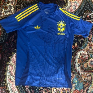  Brasilien fotbollströja - Snygg blå Brasilien fotbollströja från Adidas med gula detaljer och tre ränder på axlarna. Tryck av Kristusstatyn och stadssilhuett på framsidan. Broderad CBF-logga och Adidas Trefoil. Tillverkad i lätt och ventilerande material.