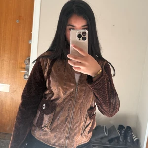 Brun jacka från Jia Yao  - Säljer en brun jacka från Jia Yao med coola manchesterdetaljer på ärmar och fickor. Jackan har dragkedja framtill, två stora fickor med knapp och blandade material som ger en unik look. 