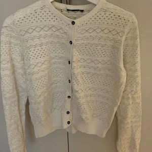 Vit stickad kofta med knappar - Superfin vit stickad kofta med snyggt hålmönster och svarta knappar framtill. Koftan har rund halsringning, långa ärmar och ribbade muddar. Perfekt att slänga över en topp för en chill och stilren look.