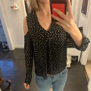 Svart prickig blus med öppna axlar - Säljer en svart blus med vita prickar och v-ringad hals. Blusen har lång ärm och cool offshoulder-detalj som visar axlarna. Materialet är lätt och luftigt, perfekt för dig som gillar att sticka ut med mönster. Passar till jeans eller kjol för en trendig look.💕