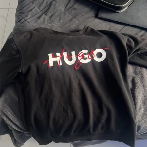 Svart sweatshirt från Hugo, storlek M - Svart sweatshirt från Hugo med stor vit och röd logotyp på bröstet. Tröjan har rund hals, ribbade muddar och är långärmad. Materialet är mjuk bomull och passformen är normal. Perfekt för dig som gillar stilrena och trendiga märkesplagg.