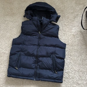 Mörkblå dunväst med huva - Snygg mörkblå dunväst med avtagbar huva och två praktiska fickor med dragkedja framtill. Västen har quiltad design och hel dragkedja framtill. Perfekt för lager-på-lager och chill stil under kyliga dagar.