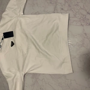 Vit sweatshirt från Prada - Säljer en stilren vit sweatshirt från Prada med klassisk rund hals och diskret logga på bröstet. Tröjan har långa ärmar och breda muddar vid ärmslut och nederkant. Perfekt för dig som gillar minimalistisk och lyxig streetstyle.