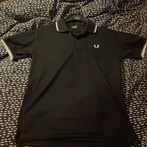 Svart piké Fred Perry M - Klassisk svart Fred Perry-piké med vit rand på krage och ärmslut. Diskret Fred Perry-logga på bröstet. Storlek: M Skick: Som ny – endast använd en gång. Snygg, tidlös modell som passar både till vardags och lite finare tillfällen.