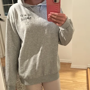 Grå hoodie med tryck 'Je m'en amour' - Grå hoodie från Gina Tricot med svart text 'Je m'en amour' och en stjärna tryckt både fram och bak. Tröjan har huva och ribbade muddar. Perfekt för en chill och avslappnad stil.