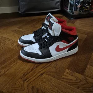 Nike Air Jordan 1 - Nike Air Jordan 1 sneakers med klassisk high-top siluett. Färgerna är svart, röd och vit med röd swoosh och detaljer. Skorna har snörning, perforerad tå och rund tå. Materialet är skinn och syntet, med röd mesh på insidan. Perfekt för dig som gillar streetwear och ikoniska sneakers.