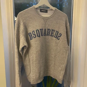 Grå sweatshirt från Dsquared2 - Snygg grå sweatshirt från Dsquared2 med blå text framtill. Klassisk rund halsringning och långa ärmar. Perfekt för dig som gillar streetwear och vill ha en stilren tröja med cool logga. Tillverkad i mjuk bomull för maximal komfort. Original pris 2000-3000 kr. Har en sliten look