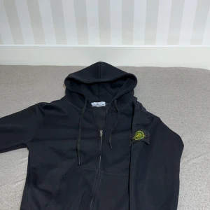 Svart hoodie från Stone Island - Svart hoodie från Stone Island med dragkedja framtill och klassisk logotyp på ärmen. Tröjan har huva med snörning och ribbade muddar. Perfekt för en avslappnad och stilren look.