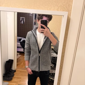 Grå stickad kofta - Blank utan märke och etiketter. Säljer en grå stickad kofta med hel dragkedja och ribbade muddar. Insidan är fodrad med rutigt tyg i svart, vitt och brunt. Koftan har hög krage och två fickor framtill. Perfekt för lager-på-lager under kyliga dagar.