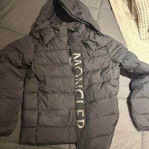  Moncler dunjacka med huva - Svart dunjacka från Moncler med stor logga längs dragkedjan och diskret Moncler-märke på ärmen. Jackan har huva, quiltad design och dragkedja framtill. Perfekt för kalla dagar och har en clean, modern look som sticker ut. Helt ny säljs för 2800 men priset går att diskutera. Storlek 2 motsvarar S/M
