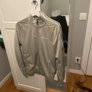 Beige vindjacka från Champion S - Snygg beige vindjacka från Champion i storlek small. Jackan har en stilren design med dragkedja framtill, ribbade muddar och två sidofickor. Perfekt för dig som gillar sportig och clean stil. Champion-logga broderad på bröstet.