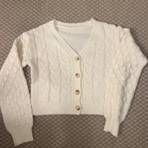 Vit stickad kofta med knappar - Mysig vit stickad kofta med grovt flätmönster och ribbade muddar. Koftan har v-ringning och stängs med fyra beige knappar framtill. Perfekt för lager-på-lager och ger en chill vibe till din outfit.