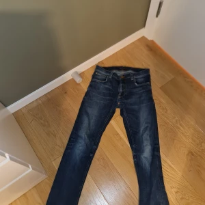 Blå jeans från Nudie Jeans Co - Snygga blå jeans från Nudie Jeans Co med klassisk femficksdesign och orangea sömmar. Jeansen har en rak passform och är tillverkade i slitstarkt denim. Perfekta för dig som gillar en tidlös och stilren look.
