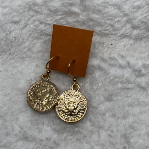 Guldiga örhängen med myntmotiv - Snygga örhängen i guldton med hängande myntformade berlocker. Mynten har detaljerade mönster och text, vilket ger en cool vintagekänsla. Perfekta för att lyfta din stil med en unik touch.