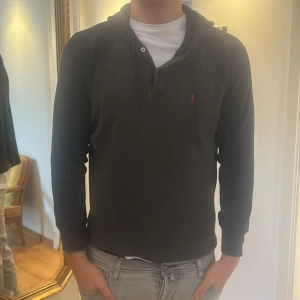 Svart hoodie med knappar och huva - Svart hoodie med diskret struktur, knappar vid halsen och huva. Liten röd detalj på bröstet. Långärmad och i mjukt material, perfekt för chill dagar. Snygg och enkel att matcha med jeans eller joggers. Skulle rekommendera denna tröja för någon som har S eller M. Jag själv bär L och den är för liten 