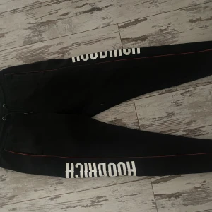 Svarta Hoodrich mjukisbyxor med tryck - Svarta mjukisbyxor från Hoodrich med vit logotyptryck längs benen och en tunn röd linje som detalj. Byxorna har snörning i midjan och sidofickor, tillverkade i mjukt material för skön passform. Perfekta för chill eller streetwear.