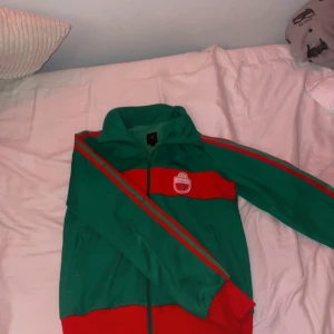 Adidas grön och röd vindjacka MAROC - Retro Adidas vindjacka i grönt och rött med MAROC-tryck på ryggen och emblem på bröstet. Jackan har dragkedja, ståkrage och klassiska ränder längs ärmarna. Perfekt för dig som vill sticka ut med sportig vintagekänsla.
