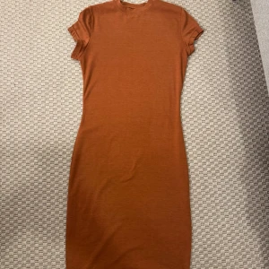 Brun kortärmad klänning i bomull - Snygg brun klänning med korta ärmar och rund hals. Modellen är figurnära och har en enkel, stilren design utan detaljer eller mönster. Perfekt för dig som gillar minimalistisk stil och mjuka material.