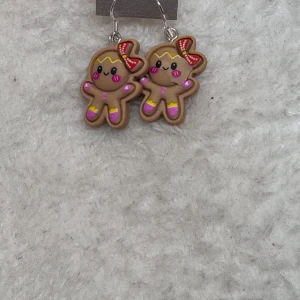 Söta pepparkaksörhängen med rosett - Unika örhängen formade som pepparkaksgubbar med glada ansikten, röda rosetter och färgglada detaljer i rosa, gul och röd. Perfekta för att ge din stil en lekfull och festlig touch. Hängande modell med silvriga krokar. .Tips till julkalendern