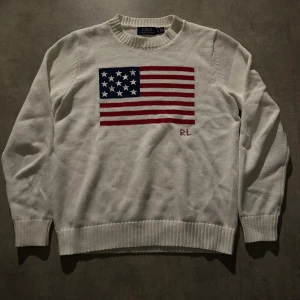 Vit stickad tröja Polo Ralph Lauren - Vit stickad tröja från Polo Ralph Lauren med amerikansk flagga i rött, blått och vitt på bröstet samt RL-broderi. Klassisk rund halsringning och ribbade muddar. Perfekt för dig som gillar preppy och tidlös stil. Hör av vid minsta fundering 💫. 