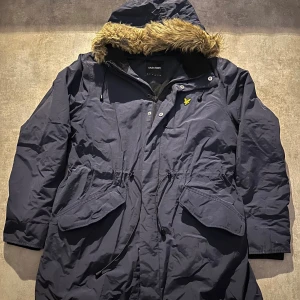 Mörkblå parkas från Lyle & Scott - Parkas i mörkblått från Lyle & Scott med huva och fuskpäls, justerbar midja med snörning och stora fickor framtill. Jackan har dragkedja och knappar samt det klassiska gula logomärket på bröstet. Perfekt för kyliga dagar. Hör av vid minsta fundering 💫