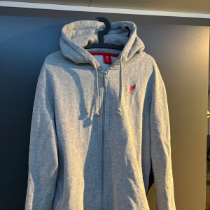Grå LFC hoodie med dragkedja - Grå hoodie från LFC i storlek L med hel dragkedja framtill och huva med snörning. Tröjan har en liten röd LFC-logga broderad på bröstet och är tillverkad i mjuk bomullsmix. Perfekt för chill dagar eller när du vill visa ditt lagstöd.