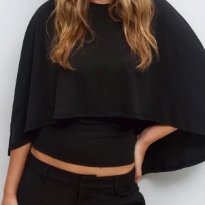 Svart cape topp XS - Svart cape topp från Gina Tricot i storlek XS. Toppens design har en cool, vid cape-liknande överdel som ger en unik siluett och croppad känsla. Materialet är mjukt och lätt, perfekt för dig som gillar statement-plagg med enkel stil. Rund halsringning och ärmlös look.