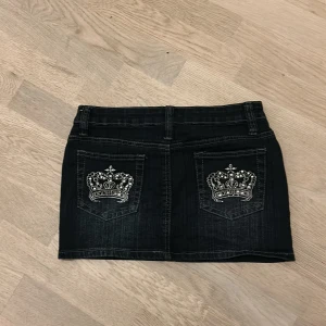 Svart jeanskjol med kronbroderi - Cool svart jeanskjol från Cotton Club med raka fickor bak, dekorerade med broderade kronor och pärldetaljer. Kjolen har klassisk femficksdesign, bälteshällor och knappstängning fram. Perfekt för dig som gillar statement-detaljer och streetstil.